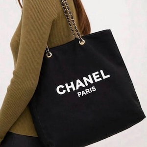 Puede incluir: Un bolso tote negro con el texto blanco CHANEL PARIS impreso en la parte frontal. El bolso cuenta con dos correas de hombro hechas de cadenas de metal dorado entrelazadas con tela negra, sujetas al bolso mediante ojales de metal dorado.