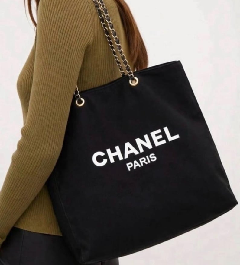 Puede incluir: Un bolso tote negro con el texto blanco CHANEL PARIS impreso en la parte frontal. El bolso cuenta con dos correas de hombro hechas de cadenas de metal dorado entrelazadas con tela negra, sujetas al bolso mediante ojales de metal dorado.