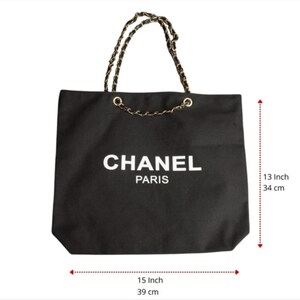 Puede incluir: Un bolso tote negro con el texto blanco CHANEL PARIS en la parte frontal. El bolso tiene asas de cadena dorada entrelazadas con material negro. Mide 34 cm de alto y 39 cm de ancho, seg&uacute;n indican las marcas de medici&oacute;n visibles.