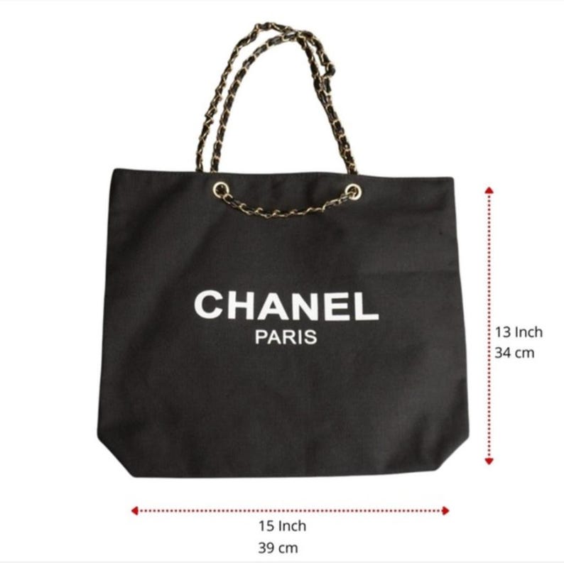 Puede incluir: Un bolso tote negro con el texto blanco CHANEL PARIS en la parte frontal. El bolso tiene asas de cadena dorada entrelazadas con material negro. Mide 34 cm de alto y 39 cm de ancho, seg&uacute;n indican las marcas de medici&oacute;n visibles.