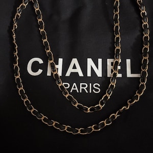 Puede incluir: Un bolso de tela negra con el texto blanco CHANEL PARIS impreso en la parte frontal. El bolso incluye una correa larga para el hombro hecha de eslabones de cadena de metal dorado entrelazados con tiras de cuero negro.