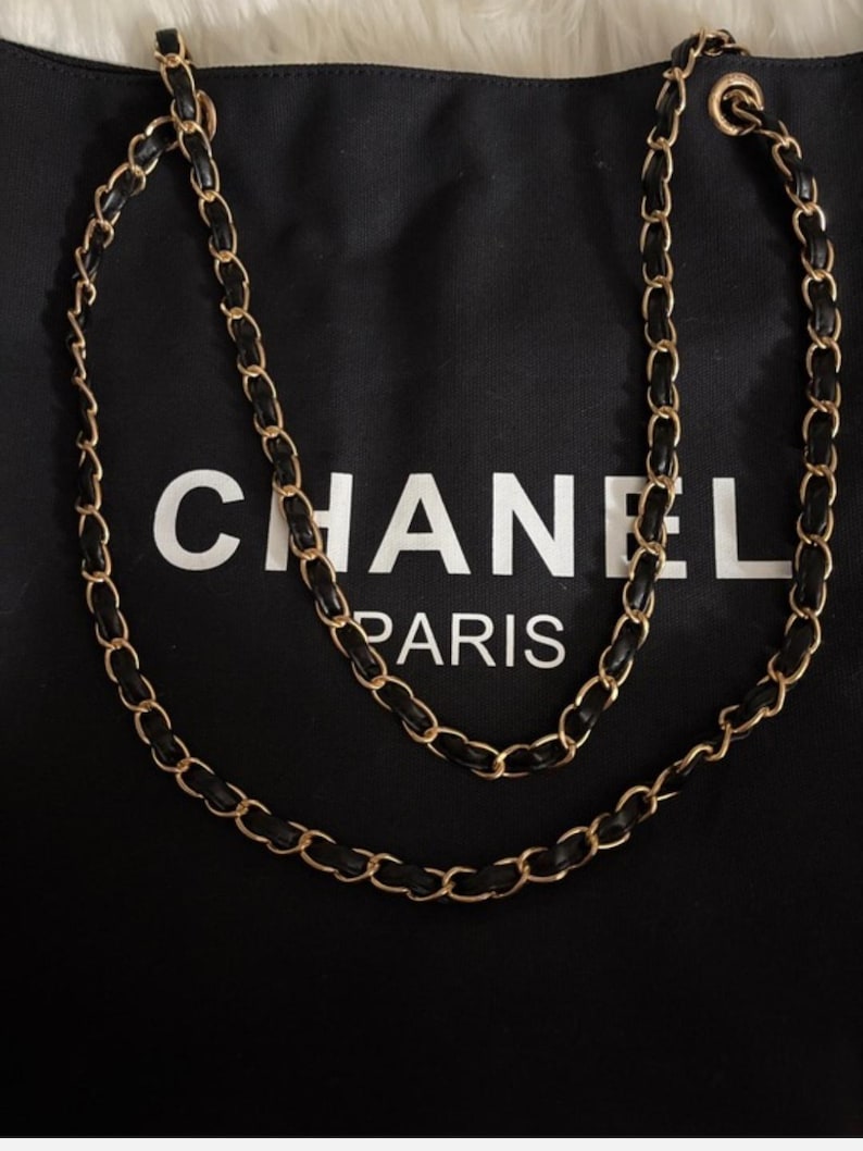Puede incluir: Un bolso de tela negra con el texto blanco CHANEL PARIS impreso en la parte frontal. El bolso incluye una correa larga para el hombro hecha de eslabones de cadena de metal dorado entrelazados con tiras de cuero negro.