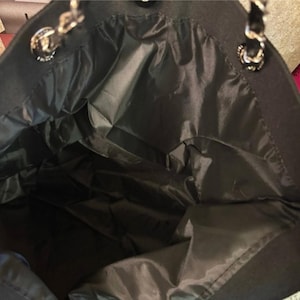 Puede incluir: Un bolso tote negro con un forro interior oscuro y liso. El bolso cuenta con dos correas para el hombro hechas de cadenas de metal plateado entrelazadas con material negro, sujetas al bolso con herrajes circulares de metal plateado.