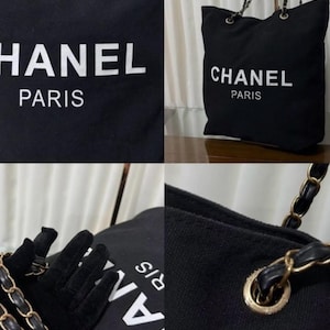 Puede incluir: Un bolso tote de lona negra con el texto blanco CHANEL PARIS impreso en la parte frontal. El bolso incluye ojales de metal dorado y una correa de cadena para el hombro entrelazada con tiras de cuero negro, ofreciendo un aspecto estructurado.