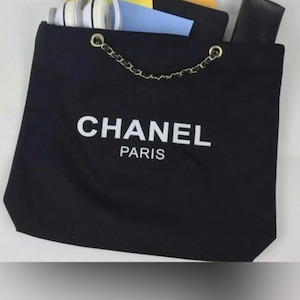 Puede incluir: Un bolso de tela negro con el texto blanco CHANEL PARIS impreso en la parte frontal. El bolso tiene un asa de cadena dorada entrelazada con material negro. Dentro hay unos auriculares blancos, un sobre azul y un estuche rectangular negro.