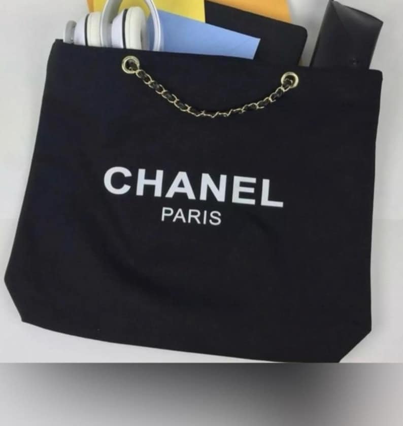 Puede incluir: Un bolso de tela negro con el texto blanco CHANEL PARIS impreso en la parte frontal. El bolso tiene un asa de cadena dorada entrelazada con material negro. Dentro hay unos auriculares blancos, un sobre azul y un estuche rectangular negro.