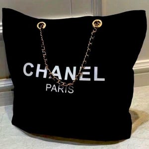 Puede incluir: Un bolso tote negro con el texto blanco CHANEL PARIS impreso en la parte frontal. El bolso incluye dos asas de cadena de metal dorado entrelazadas con material negro y sujetas a la parte superior con anillas de metal dorado.