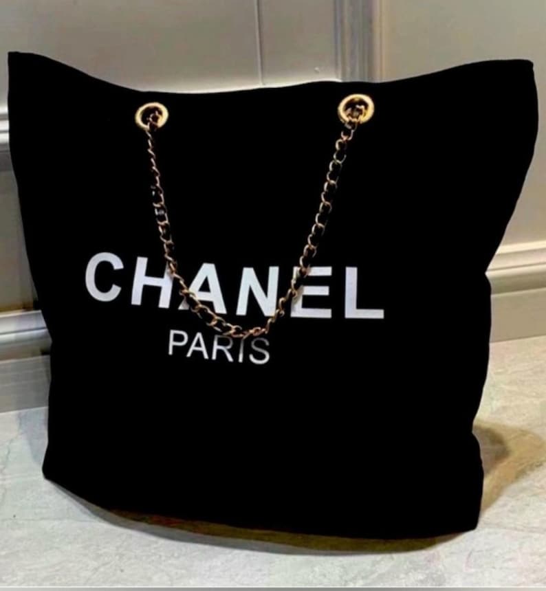 Puede incluir: Un bolso tote negro con el texto blanco CHANEL PARIS impreso en la parte frontal. El bolso incluye dos asas de cadena de metal dorado entrelazadas con material negro y sujetas a la parte superior con anillas de metal dorado.