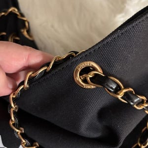 Puede incluir: Un bolso de mano de lona negra con una correa de cadena de metal dorado entrelazada con cuero negro. La correa pasa a trav&eacute;s de un ojal circular de metal dorado grabado con la palabra CHANEL. El bolso tiene un exterior de tela texturizada.