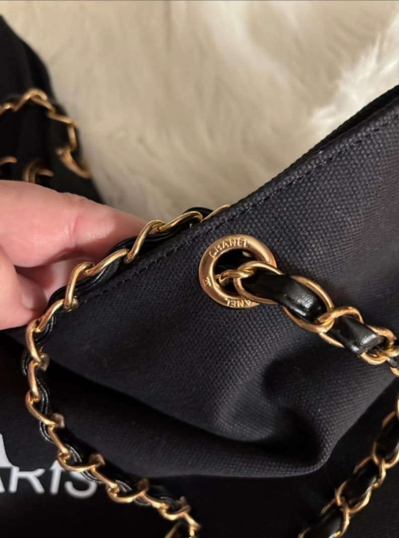 Puede incluir: Un bolso de mano de lona negra con una correa de cadena de metal dorado entrelazada con cuero negro. La correa pasa a trav&eacute;s de un ojal circular de metal dorado grabado con la palabra CHANEL. El bolso tiene un exterior de tela texturizada.