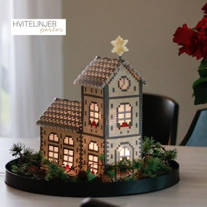 May include: A decorative, illuminated house made of small, light-colored beads, with a star on top. The house has multiple windows that glow with warm light, and is surrounded by greenery on a black tray. The text "HVITELINJER perler" is in the upper left corner.
