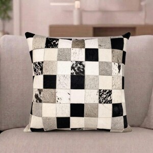 Könnte beinhalten: Ein quadratisches Zierkissen mit einem Patchwork-Design aus abwechselnden Quadraten in Schwarz, Weiß und verschiedenen Grautönen. Die Oberfläche besteht aus einzelnen texturierten Flicken, die in einem Raster angeordnet sind und ein Schachbrettmuster bilden.