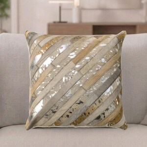 May include: A square throw pillow featuring a patchwork design of diagonal strips in varying shades of tan, beige, gray, and metallic silver. The surface displays a mix of textured hair-on-hide patterns and smooth, reflective metallic finishes.