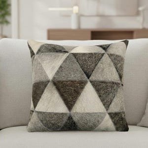 May include: Square decorative pillow with a geometric pattern of triangles in shades of gray and brown. The pillow has a textured, fur-like appearance and is placed on a light-colored sofa. The pillow is approximately 18 inches square.