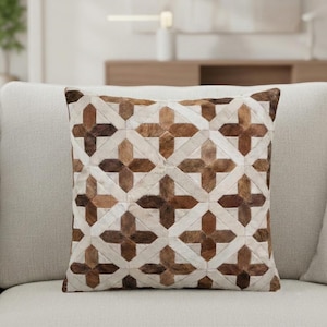 May include: Square cushion with a geometric pattern. The cushion features a patchwork design with brown and cream-coloured leather pieces arranged in a cross pattern. The cushion is placed on a light-coloured sofa.
