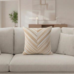 Puede incluir: Un cojín decorativo con un patrón de chevron en tonos beige y crema descansa sobre un sofá beige. El diseño del cojín presenta rayas diagonales, creando una apariencia texturizada. El sofá forma parte de un salón de tonos neutros. El cojín mide aproximadamente 40 cm x 40 cm.