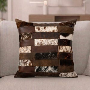 May include: A square throw cushion featuring a patchwork design of alternating rectangular panels. The pattern consists of dark brown leather strips and textured cowhide sections in shades of white, brown, and black with natural hair patterns.
