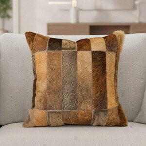 May include: A square throw pillow constructed from rectangular patches of genuine cowhide leather. The pillow features a patchwork design with varying shades of brown, tan, and dark chocolate fur, arranged in a vertical and horizontal grid pattern.