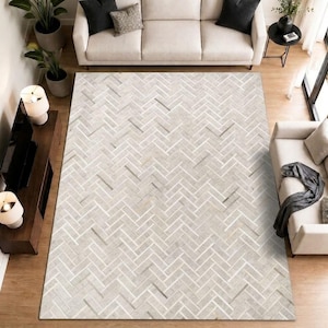 Puede incluir: Una alfombra rectangular grande con un patrón de espiga en gris claro y blanco. La alfombra está compuesta por segmentos rectangulares individuales dispuestos en un diseño de zigzag repetitivo, cubriendo el espacio del suelo entre dos sofás.