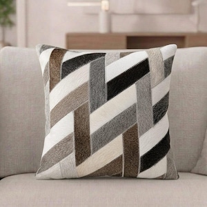 Può includere: Un cuscino quadrato con un motivo geometrico a strisce diagonali nei toni del bianco, grigio, marrone e nero. La superficie ha un aspetto testurizzato simile alla pelle con cuciture visibili lungo i bordi di ogni singola striscia.