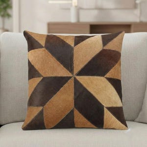 Könnte beinhalten: Quadratisches Zierkissen mit geometrischem Muster. Das Kissen hat ein Patchwork-Design aus braunen und beigefarbenen Lederstücken, die in einem Sternmuster angeordnet sind. Das Kissen liegt auf einem hellen Sofa.