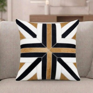 May include: A square throw cushion featuring a geometric starburst pattern composed of alternating strips of black, white, and tan textured material. The panels are joined with visible stitching, creating a symmetrical design that radiates from the centre.