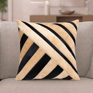 May include: A square throw pillow featuring a geometric pattern of alternating diagonal stripes in black and tan. The surface of the pillow is constructed from panels of hair-on hide, creating a textured appearance with distinct color blocks.