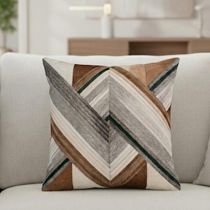 Puede incluir: Un cojín cuadrado con un diseño geométrico de patchwork hecho de cuero con pelo. El patrón consiste en rayas diagonales alternas en tonos crema, bronceado, marrón oscuro, gris y negro, creando un efecto de chevrón simétrico en la superficie.