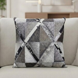 May include: Square decorative pillow with a patchwork design. The pillow features a geometric pattern of various shades of gray, black, and white, creating a textured appearance. The pillow is placed on a light-colored sofa.