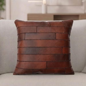 May include: A square throw pillow constructed from multiple rectangular pieces of brown leather stitched together. The leather features a natural grain texture with varying shades of dark and medium brown, arranged in a horizontal brick-like pattern.