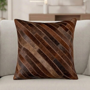 Puede incluir: Un cojín cuadrado con un diseño de patchwork de tiras diagonales en varios tonos de marrón. La superficie está compuesta de piel animal texturizada con un acabado natural de pelo, creando una apariencia de múltiples tonos en el frente.