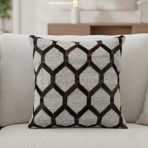 May include: Square cushion with a geometric pattern. The cushion has a light grey background with a dark brown honeycomb design. The cushion is placed on a light-coloured sofa, suggesting a home decor theme.