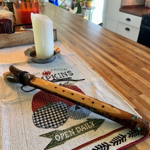 Puede incluir: Una flauta de madera hecha a mano con un aspecto natural y rústico. La flauta presenta agujeros para los dedos y adornos turquesas. Descansa sobre una tela estampada con las palabras "FRESH PUMPKINS" y "OPEN DAILY".