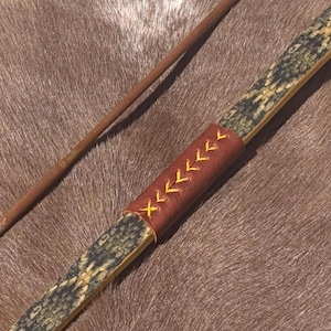 May include: A handcrafted bow with a snakeskin pattern and a brown leather grip stitched with yellow thread. A wooden arrow rests nearby, suggesting archery or hunting equipment.