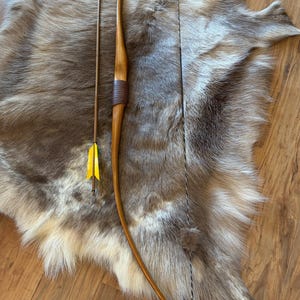 以下が含まれることがあります： 茶色の革のグリップが付いた伝統的な木製の弓と、黄色の羽根が付いた1本の矢。 弓と矢は、木製の床を背景に、質感のあるライトブラウンと白の動物の毛皮のラグの上に展示されています。