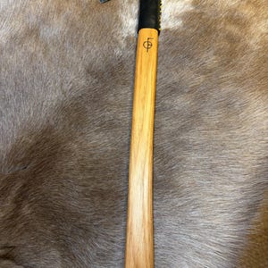 Puede incluir: Un tomahawk con mango de madera y cabeza negra. El mango es de color marrón claro con un símbolo grabado. La cabeza es negra con una pequeña hoja de hacha. El mango está envuelto en cuero negro cerca de la cabeza.