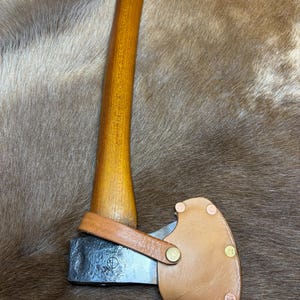 May include: A small axe with a wooden handle and a leather sheath. The handle is a light brown color, and the axe head is silver. The leather sheath is tan and secured with a leather strap and brass rivets.