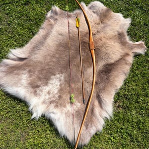 Puede incluir: Un arco tradicional de madera con empuñadura de cuero y una sola flecha con plumas amarillas y verdes, apoyado sobre una piel de animal marrón claro. El arco está curvado y la flecha está lista para ser disparada. El fondo es hierba verde.