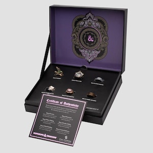Puede incluir: Una caja negra que contiene siete anillos de fantasía, cada uno con diseños y colores únicos. La caja tiene un interior púrpura con un emblema decorativo. Se incluye un certificado de autenticidad, con el texto "Dungeons & Dragons".
