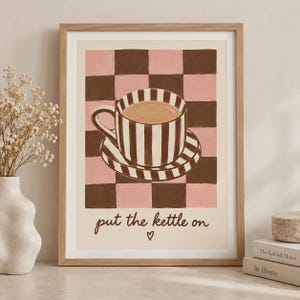 May include: A framed wall art print featuring a brown and white striped teacup on a matching saucer, centred over a pink and brown checkered background. Below the cup, the text reads "put the kettle on" with a small heart symbol underneath.