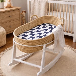 May include: A woven baby bassinet on a white rocking frame. The bassinet features a blue and white patterned sheet and a white blanket. The room has a neutral colour scheme with a round jute rug.