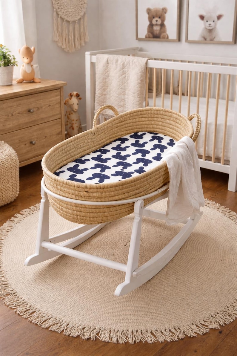 May include: A woven baby bassinet on a white rocking frame. The bassinet features a blue and white patterned sheet and a white blanket. The room has a neutral colour scheme with a round jute rug.