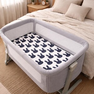 May include: A grey and white baby bassinet with a navy blue bunny pattern on the mattress. The bassinet is next to a bed with beige bedding. The room has a neutral colour palette.