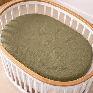 Può includere: Un lenzuolo con angoli verde oliva all'interno di una culla ovale con barre bianche e una struttura in legno. Il lenzuolo ha un aspetto strutturato e stropicciato. La culla è progettata per il comfort del bambino.