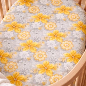 Puede incluir: Una s&aacute;bana bajera para cuna con un estampado floral. La s&aacute;bana presenta grandes flores amarillas con centros naranjas y flores blancas m&aacute;s peque&ntilde;as sobre un fondo gris. La s&aacute;bana est&aacute; dentro de una cuna de madera.