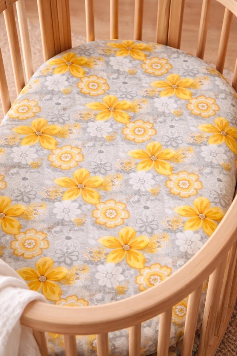 Puede incluir: Una s&aacute;bana bajera para cuna con un estampado floral. La s&aacute;bana presenta grandes flores amarillas con centros naranjas y flores blancas m&aacute;s peque&ntilde;as sobre un fondo gris. La s&aacute;bana est&aacute; dentro de una cuna de madera.