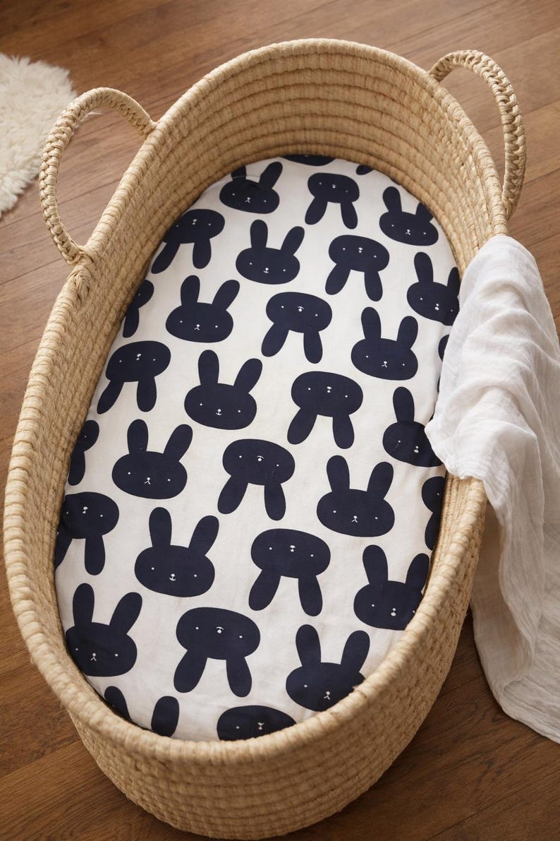 May include: A wicker bassinet with a white sheet patterned with navy blue bunny faces. A white muslin blanket is draped over the side. The bassinet has handles and sits on a wooden floor.