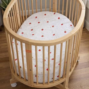 May include: A light wood oval crib with vertical slats and a mattress featuring a strawberry pattern. The crib has wheels for easy movement. The brand name "Stokke" is visible on the mattress.
