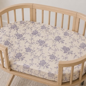 May include: A light wood oval baby bassinet with a floral patterned sheet. The sheet features a cream background with a repeating pattern of purple roses and vines. The bassinet has a slatted design.