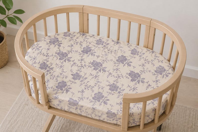 May include: A light wood oval baby bassinet with a floral patterned sheet. The sheet features a cream background with a repeating pattern of purple roses and vines. The bassinet has a slatted design.
